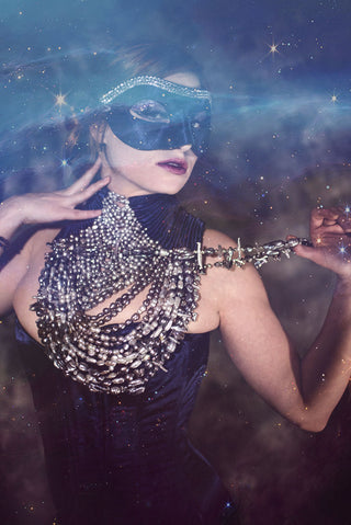 A woman in a navy embellished venetian mask wears a deep blue corset-ed dress with a high neckline with detailed pearl neck-piece. The background is galaxy themed.
