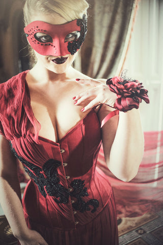 A blonde woman in a red mask stands in an ornate parlour. She wears a deep red corset dress with detailed black beading and floral elements.